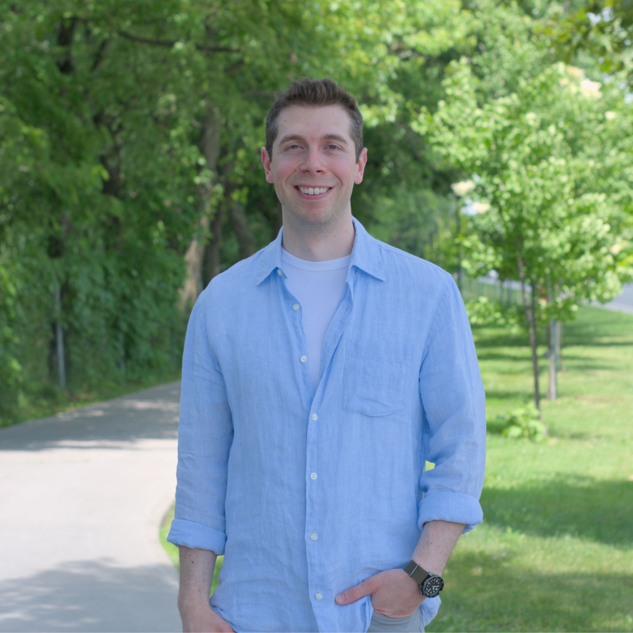 Pierre-Luc Brodeur, candidat dans le District 2 : Un homme souriant, aux cheveux courts, porte une chemise bleu pâle par-dessus un t-shirt blanc. Il est debout sur un sentier bordé d’arbres, une main dans la poche, par une journée ensoleillée. L’arrière-plan montre une rangée d’arbres verts et une route