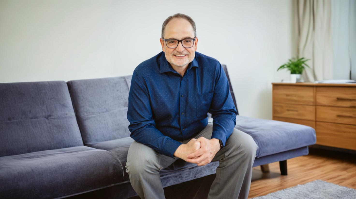 Photo de Daniel Picard, Candidat à la mairie : Un homme d'âge moyen, souriant, avec une barbe grise et des lunettes, est assis sur un canapé gris dans un salon lumineux. Il porte une chemise bleue et un pantalon gris pâle. À l’arrière-plan, on voit une commode en bois, une plante verte et un rideau beige