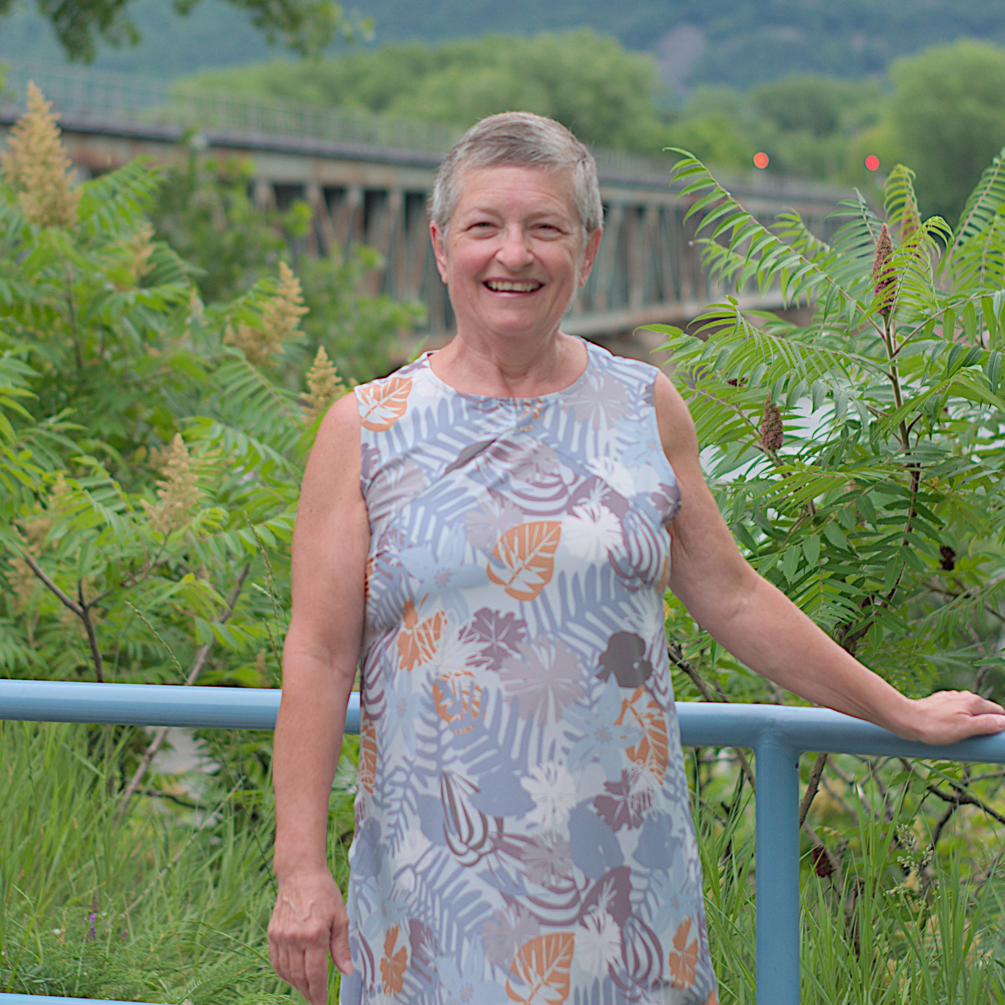 Louise Allie, candidate dans le District 1 :  Une femme souriante aux cheveux gris courts pose à l’extérieur, vêtue d’une robe sans manches à motifs floraux et feuillus dans des tons pastel. Elle est appuyée sur une rambarde bleue, entourée de végétation verdoyante. À l’arrière-plan, on aperçoit un pont métallique ainsi que le Mont-Saint-Hilaire.