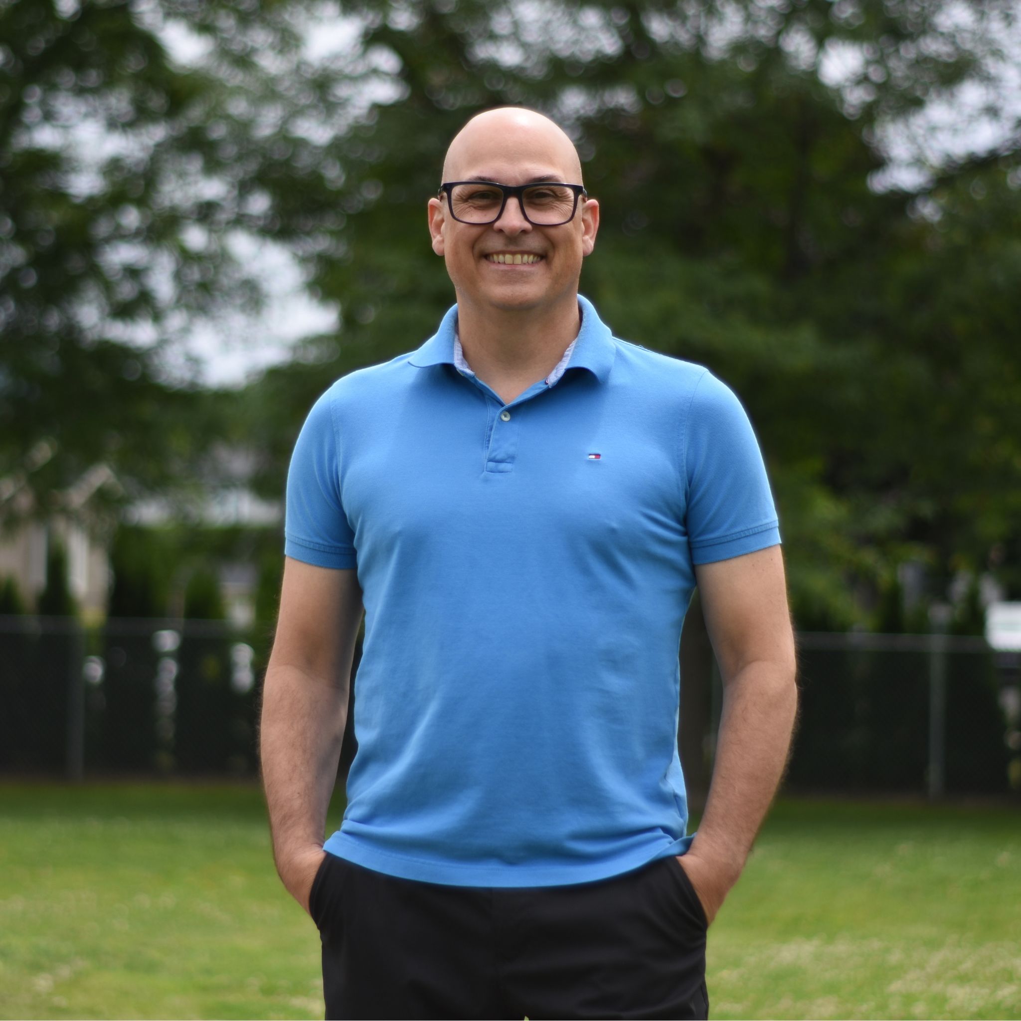 Vincent Chabot, candidat dans le District 4 : Un homme souriant, chauve et portant des lunettes, pose debout à l’extérieur. Il est vêtu d’un polo bleu et d’un pantalon foncé, les mains dans les poches. Derrière lui, on voit une clôture grillagée, une pelouse et des arbres.