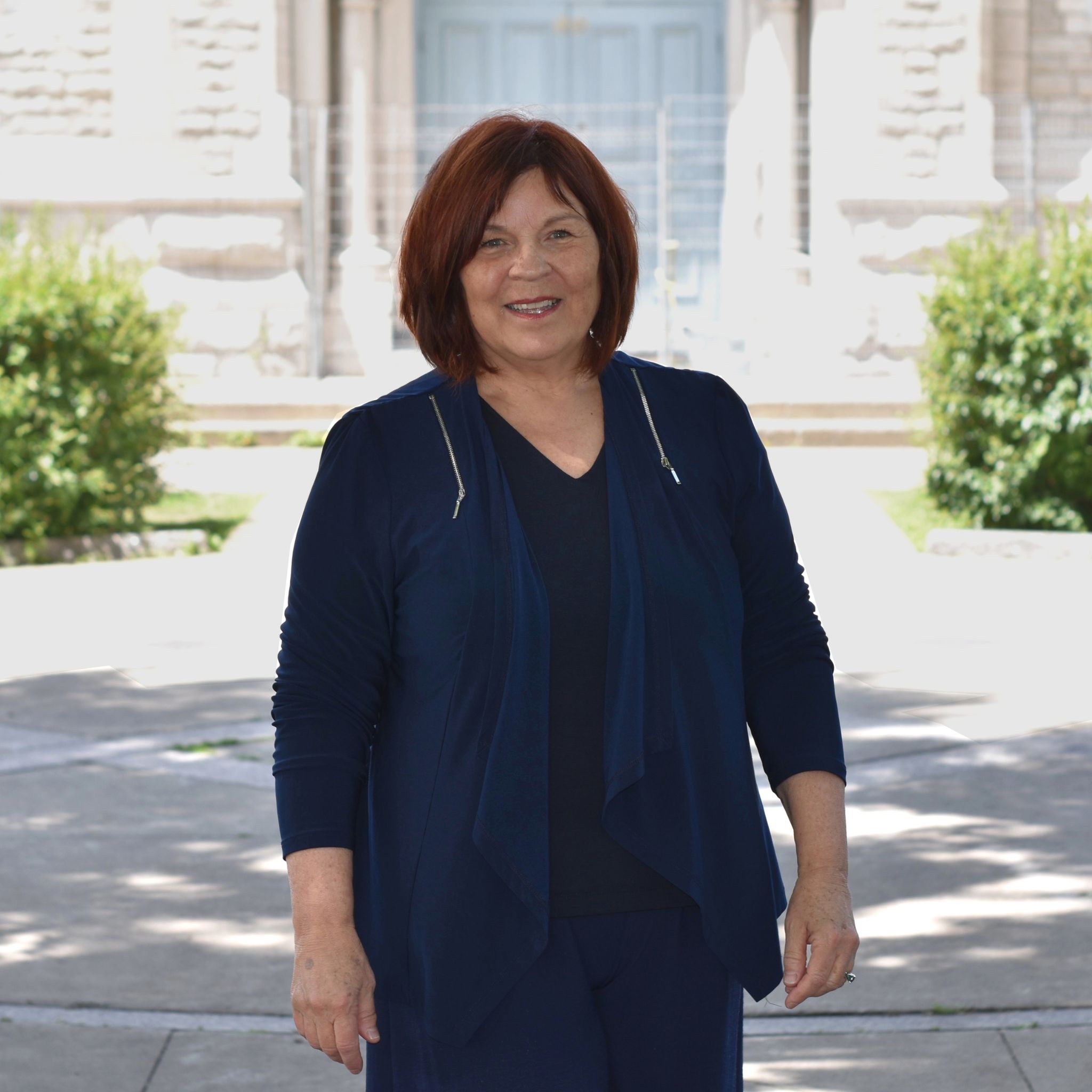 Hélène Gosselin, candidate dans le district 5 :  Une femme souriante aux cheveux auburn mi-longs se tient debout à l’extérieur devant un bâtiment de pierre clair. Elle porte un ensemble bleu foncé avec un haut fluide et un pantalon assorti. À l’arrière-plan, on distingue des escaliers, deux arbustes et une grande porte bleue pâle.