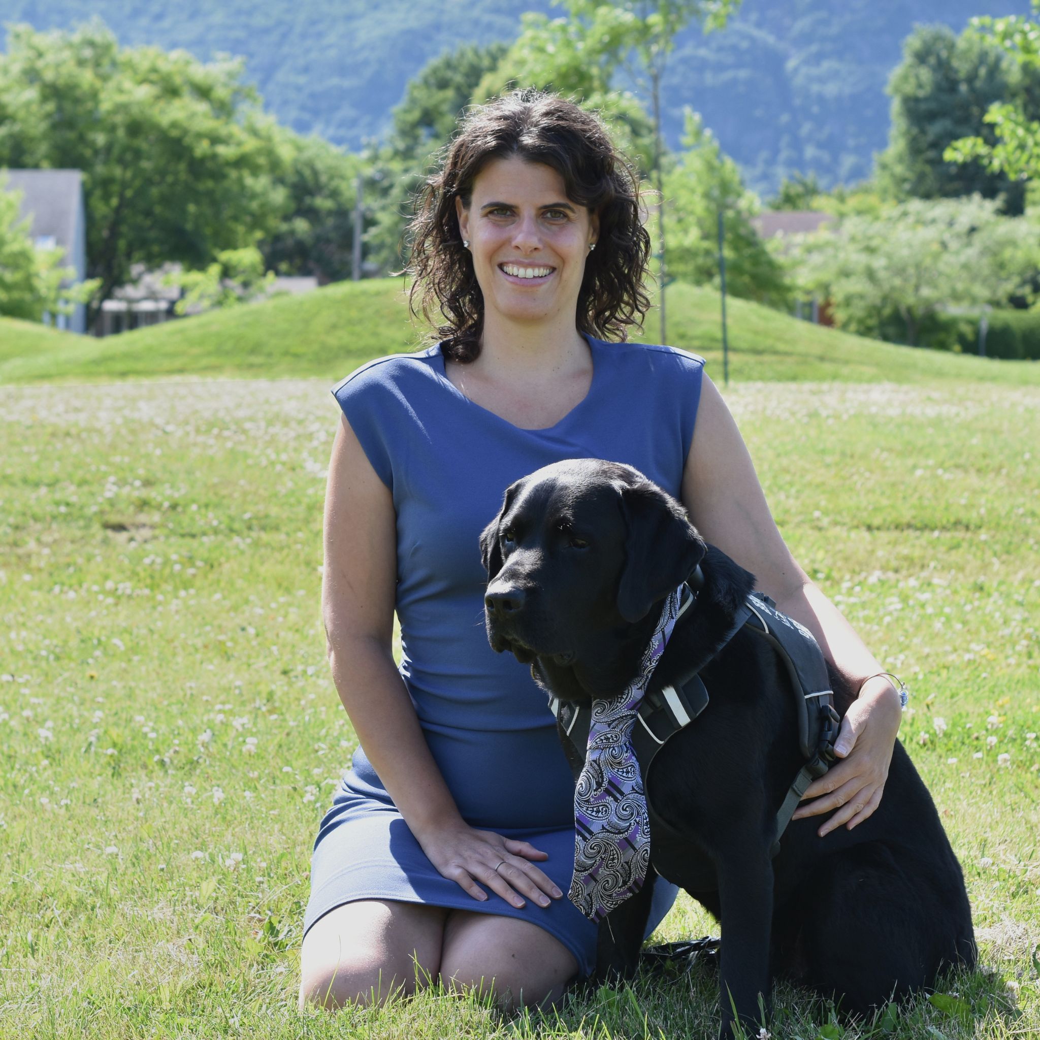 Claudia Nigrelli, candidate dans le District 6 : Une femme souriante, aux cheveux bruns bouclés, est agenouillée sur l’herbe dans un parc verdoyant, avec des collines et des arbres en arrière-plan. Elle porte une robe bleu mauve et tient un chien guide noir assis à ses côtés, qui porte un harnais et une cravate décorative. 