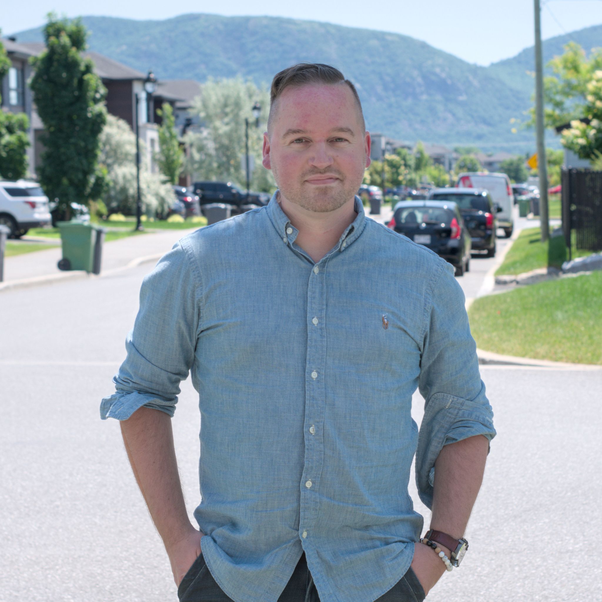 Youann Blouin, candidat dans le District 7 :  Un homme debout dans une rue résidentielle, les mains dans les poches, regarde la caméra avec un léger sourire. Il porte une chemise bleu pâle à manches retroussées. En arrière-plan, on voit des maisons, des voitures stationnées et une montagne verdoyante sous un ciel clair. 